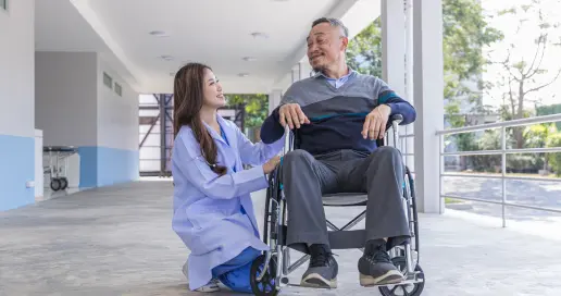 A healthcare worker kneels beside an elderly man seated in a wheelchair. They are outside in a hallway with natural light, and both of them are smiling and engaged in conversation. The setting appears to be a care facility or hospital.