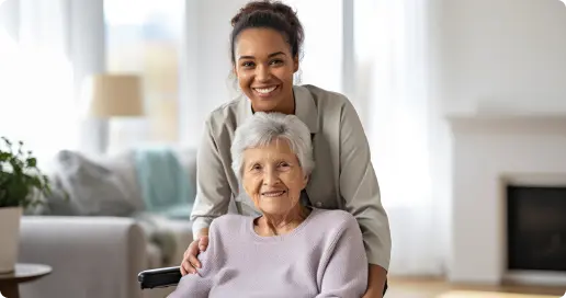 A smiling elderly woman sits in a wheelchair inside a well-lit living room. Behind her stands a younger woman, also smiling, with her hands gently placed on the elderly woman's shoulders. The room features a cozy and modern decor with a plant and soft furnishings.