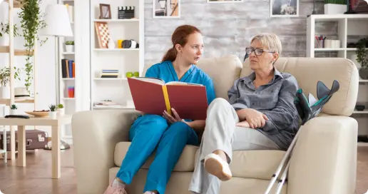 A caretaker in blue scrubs is sitting on a couch with an older woman holding a red book. The elderly woman, wearing glasses and a grey outfit, has crutches resting beside her while they have a conversation in a well-lit living room with various decorations and bookshelves.