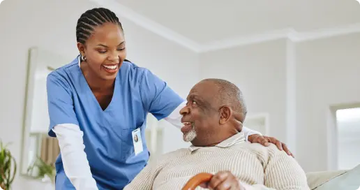 A nurse in blue scrubs smiles while interacting with an elderly man who is sitting and holding a cane. The nurse has her hand on the man's shoulder, and they appear to be having a pleasant conversation in a bright, homey environment.